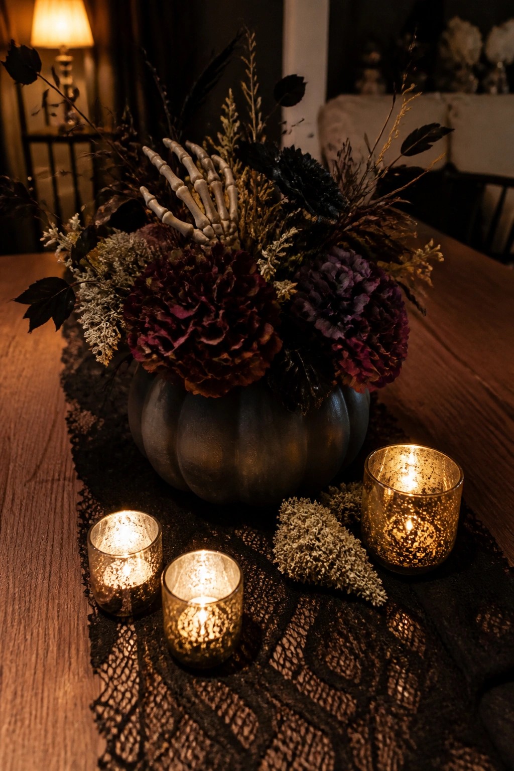 Dark pumpkin vase with purple flowers and skeleton hand amid lit gold candles on lace runner