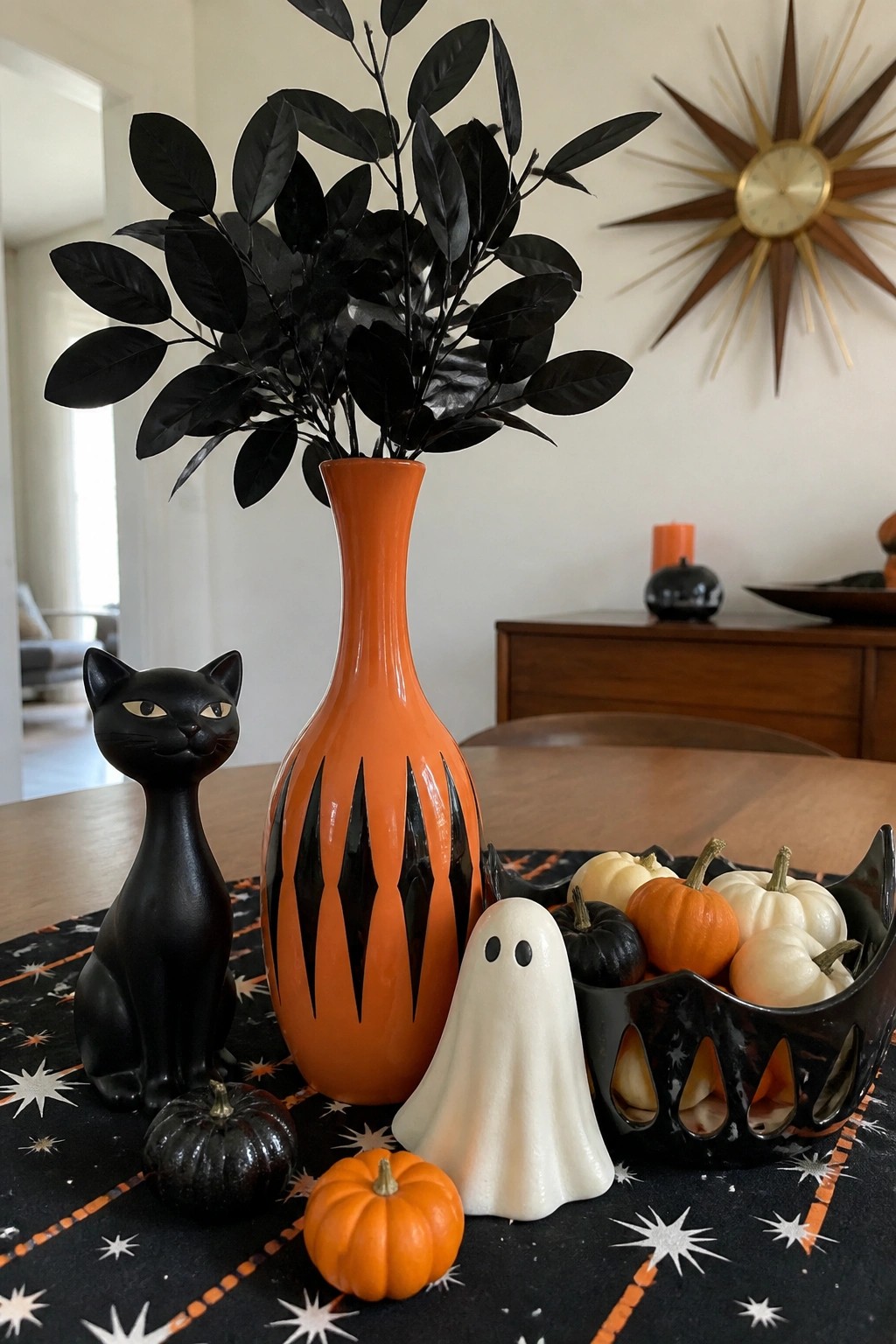 A dining table centerpiece with an orange vase holding black foliage, a black cat figurine, a white ghost, and small pumpkins arranged on a black patterned tablecloth.