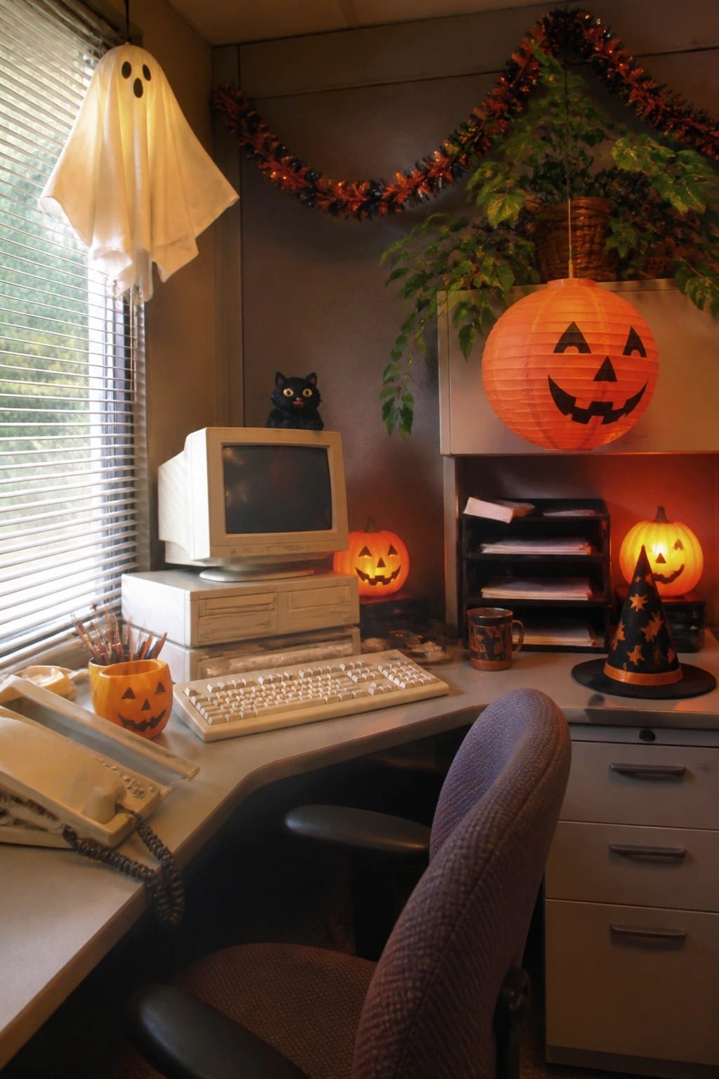 Office desk with a hanging white sheet ghost, multiple jack o lanterns, a black cat figurine, and a vintage computer setup.