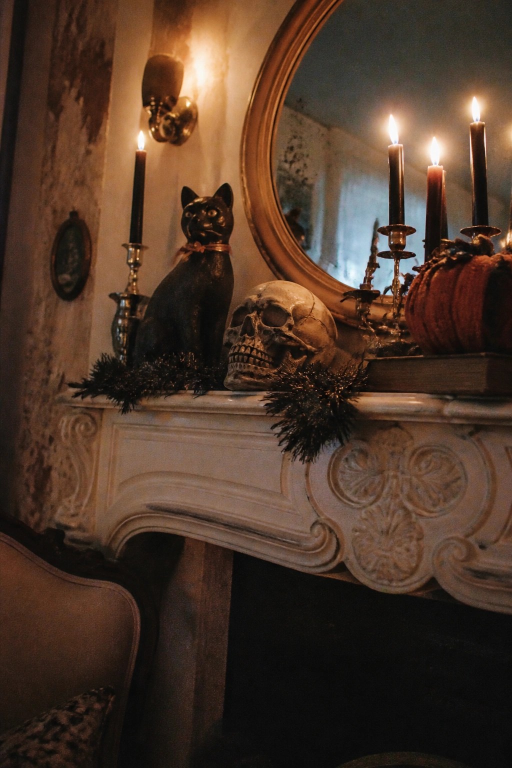 White ornate fireplace mantel with a black cat statue, human skull, lit black candles, orange pumpkin, and dark garland.