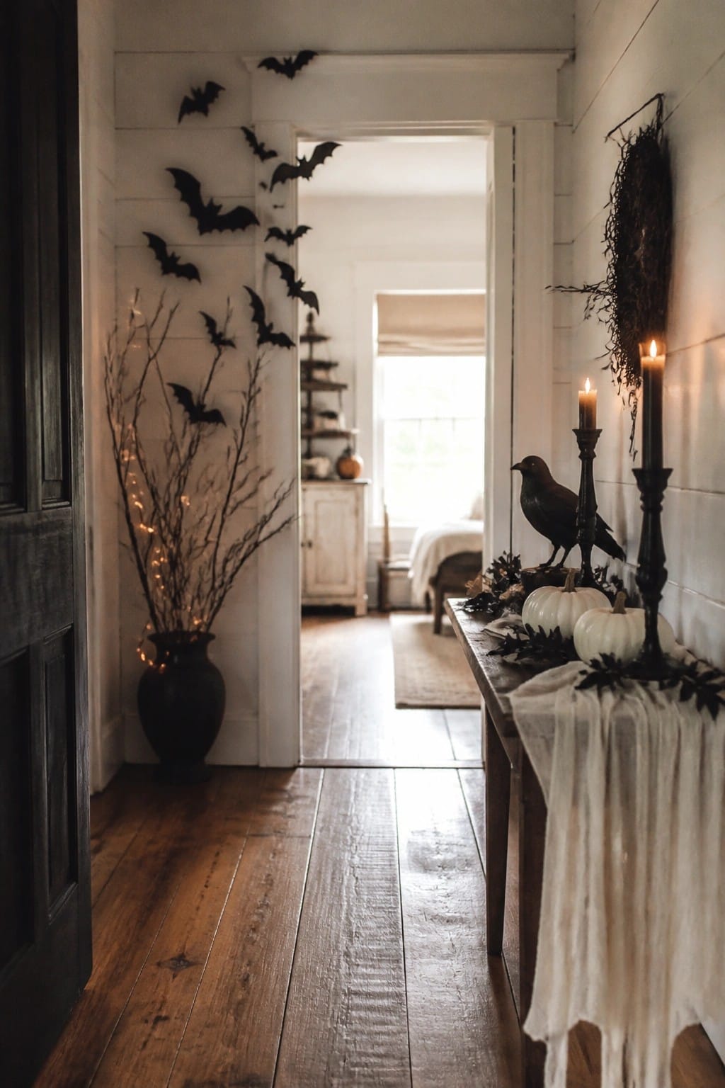 White entryway hallway with black bat cutouts arranged on the wall beside a wooden console holding a black crow, white pumpkins, and lit black candles.